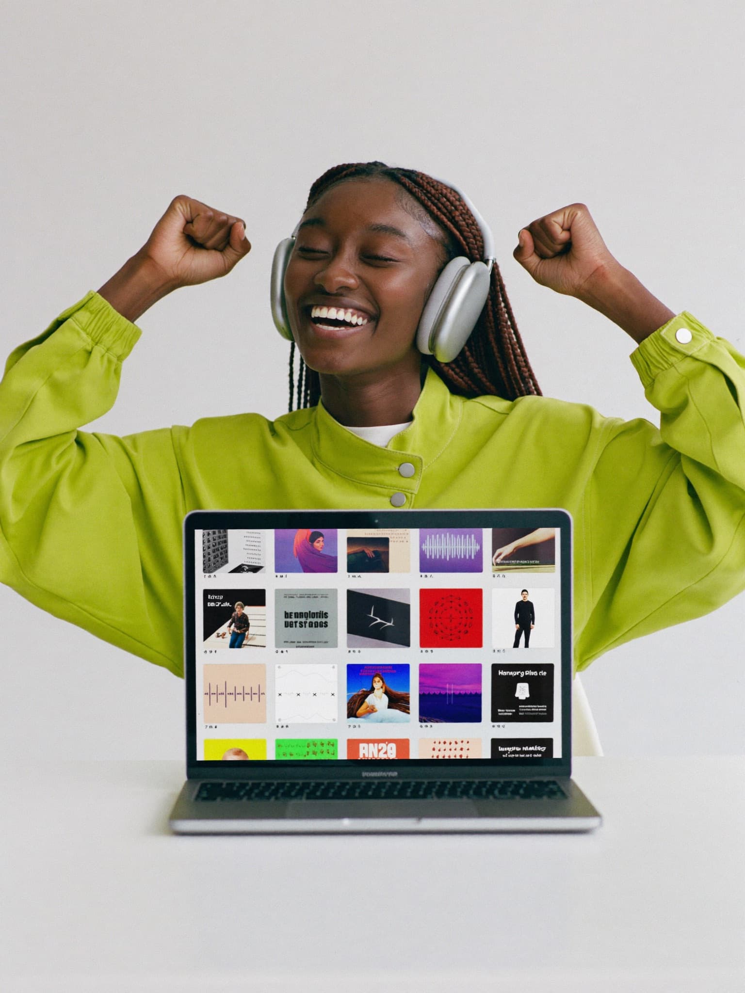 A music artist in headphones laughing with both fists raised in celebration at a bright white studio desk, her organized music catalog on the laptop in front of her — her song just landed a placement.