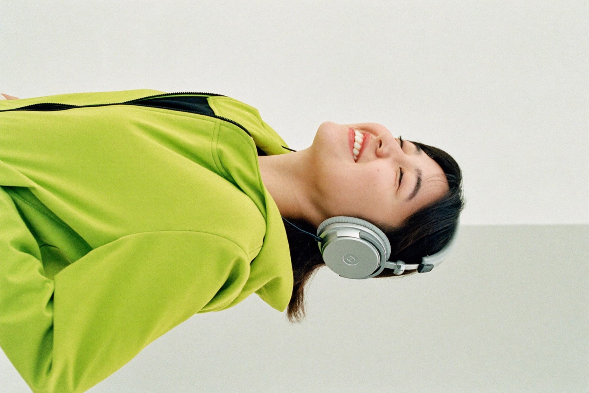 A young artist in a lime-green jacket and headphones, eyes closed, smiling as they nod along to music.